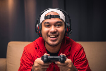 Playing video game. Young asian handsome man sitting on sofa holding joystick in living room. Happiness Streamer Indian man wearing headset playing game online in the darkroom.