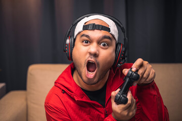 Excited and shocked face of Asian gamer with headphone holding headset playing video game online sitting on sofa at living room. Indian professional gamer streaming on social playing game very fun © Chanakon