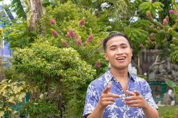 A confident Filipino guy in a hawaiian shirt doing the finger gun gesture. Outdoor scene.