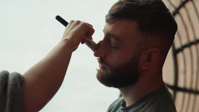 Close-up hand of professional make up artist putting tone powder on young bearded male model face at beauty studio salon