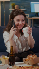 Caucasian adult enjoying phone call with friend in living room while drinking coffee. Woman talking on smartphone, preparing to eat fast food delivery meal at home after work to relax