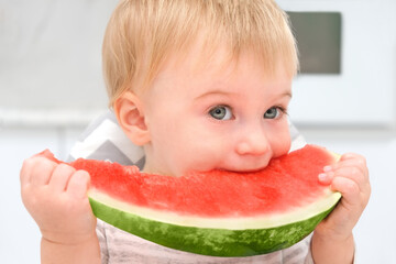 Cute caucasian blonde baby 1 year old,having lunch at kitchen,eating red water melon.Healthy fresh fruit and berry for infant concept.Child having tasty meal.Kid and food concept.Close-up.