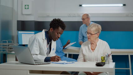 Caucasian aged patient receiving consultation from doctor in medical office at facility. Medic examining disease symptoms for sick older woman while nurse discussing with senior man