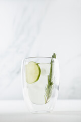Detox cold lime lemonade with ice cubes, lime slices, rosemary twig in transparent mug on white wood table in sunlight, summer cocktail, vertical.