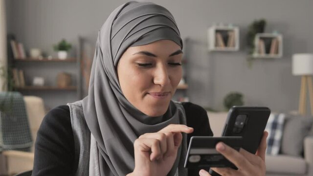 Happy muslim arab woman wearing hijab holding smart phone mobile and credit card shopping online,satisfied middle eastern girl in a modern apartment makes payment on internet