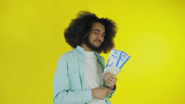 A happy African-American man looks at the camera, holds tourist vouchers in his hand, stands isolated on a yellow background