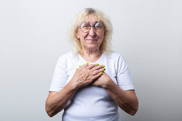 Smiling old woman with glasses holds her hands on her chest on a light background