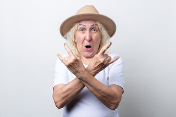 Surprised old woman in hat shows rocker horn gesture on light background