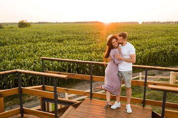 Beautiful joyful couple standing at the field, hugging, having a date in sunset, enjoying moments together. Handsome man gently cuddling and kissing adorable smiling woman, weekends outdoors concept