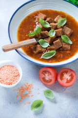 Tomato and lentil soup topped with croutons and fresh green basil, vertical shot on a light-blue stone surface, close-up