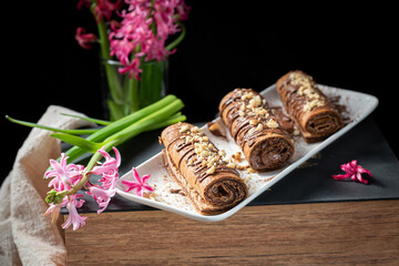 Chocolate roll with chocolate cream with nuts. Chocolate cakes on a white plate, decorated with flowers on a black background.
