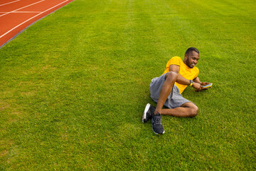 Fototapeta na wymiar Happy young adult man laying on the grass, using smartphone, browsing internet. Joyful african american male relaxing, taking a break after workout outdoors, having a video call from friends, smiling