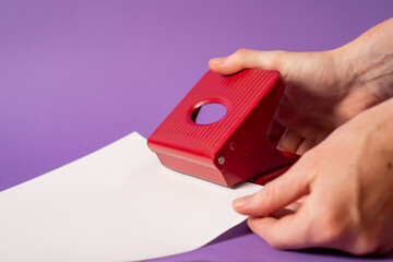 Hand holding paper hole puncher on violet background. Red office paper hole punch. The girl makes paper holes with punch