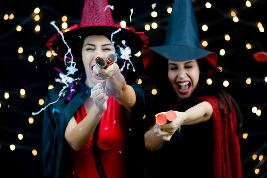 Shiny glossy paper confetti popper cracker exploded when pulled by two happy funny excited female models in black red witch dress costume tall hat and shawl in blurred background in Halloween party