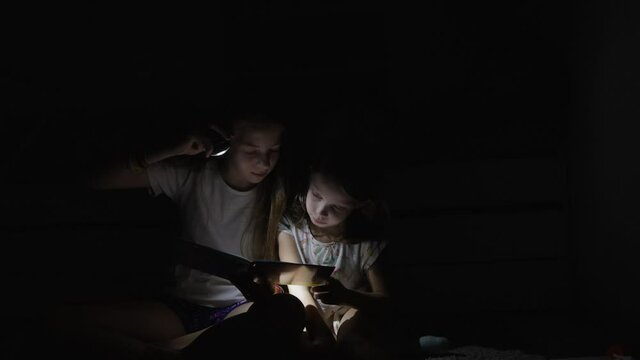 two sisters reading a book at night with a flashlight under the covers