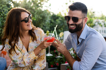 Cheerful couple of young people having fun laughing clinking tropical cocktails at a garden terrace party. Friends toasting outdoors.