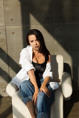 Obraz na płótnie Canvas Pretty young woman with big breast posing on white chair in studio, steel wall behind, wearing jeans and white shirt, natural warm sunset light
