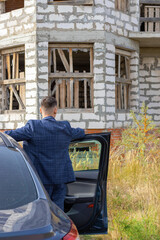 young man is standing near the car in front of an unfinished house, construction concept or mortgage crisis, inability to pay for housing