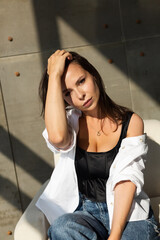 Fototapeta premium Pretty young woman with big breast posing on white chair in studio, steel wall behind, wearing jeans and white shirt, natural warm sunset light