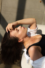Pretty young woman with big breast posing on white chair in studio, steel wall behind, wearing jeans and white shirt, natural warm sunset light