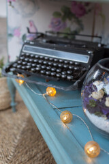 garland of led lights with golden spheres next to an old typewriter and a glass container with colored flowers on a blue wooden table on a brown carpet - vertical - ornaments - events - weddings