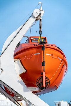 Vertical shot of orange life boat hanging on a crane onboard sailing ocean ship with blue sky in the back.