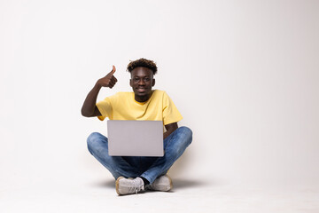 Excited african man showing thumbs up while sitting with laptop on floor over white background