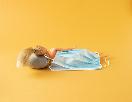 A doll lies down and sleeps on a grey pillow covered with a mask on yellow background. Minimal covid 19 concept.