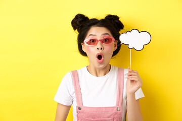 Summer and fashion concept. Excited party girl in sunglasses, holding comment cloud party mask and gasping amazed, standing impressed on yellow background