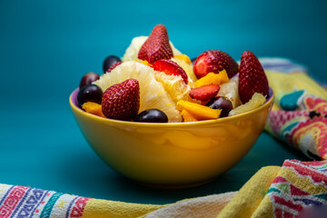 Uma porção de frutas dentro de uma vasilha amarela com fundo na cor azul tiffany.