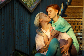 Lesbian loving young family in casual clothes sits hugging on old rusty steps in city park at sunset light in summer evening