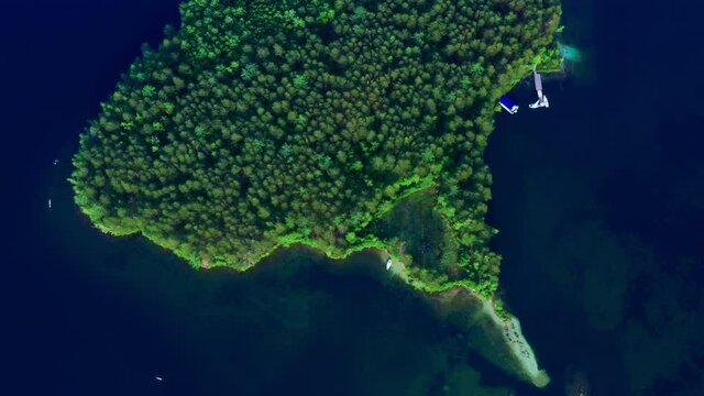 Small beautiful island in ocean covered with green plants, trees taken by drone. Aerial, top view of river ait, eyot, white yachts, seagulls flying over it. Sea isle, blue water surface
