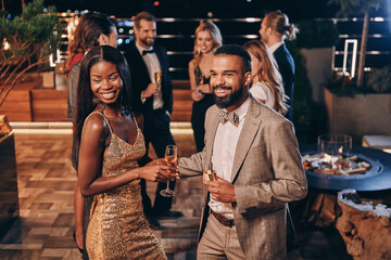 Beautiful young African couple in formalwear holding champagne flutes and smiling - obrazy, fototapety, plakaty