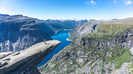 Fototapeta na wymiar Girl on top of the Troll Tongue in Norway cliff facing blue fjords and snow capped mountains