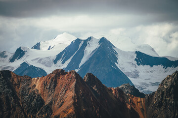 Atmospheric mountains landscape with great snowy top behind colorful brown red orange rocky wall under cloudy sky. Dramatic highland scenery with giant glacier and big vivid brown red orange mountain.