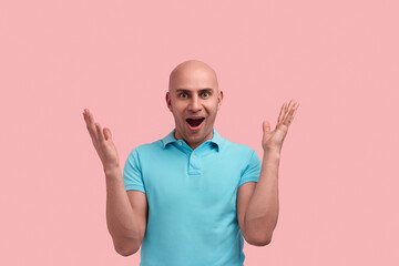 Fototapeta premium Happy bald homosexual man with bristle is positively surprised by product size, shows big object with hands, opens mouth in excitement, gay friendly, wears blue polo shirt, poses over pink background.