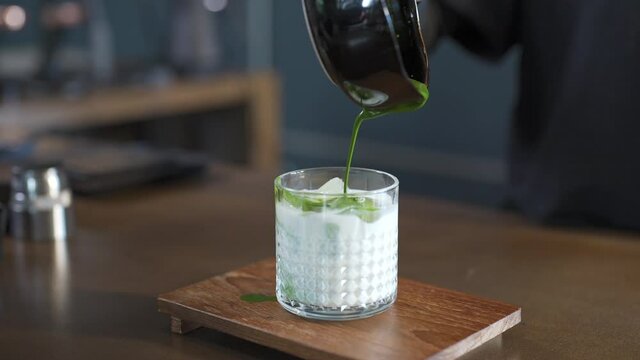 4K Asian woman barista pouring green tea on fresh milk in the glass. Female coffee shop waitress serving iced matcha latte to customer on counter at cafe. Small business coffee shop owner concept.