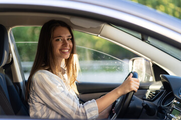 Fototapeta na wymiar Happy young girl sitting on driver seat in new car joyful smiling hold hands on wheel. Cheerful female driving vehicle looking through open window. Successful woman car owner or getting driver license
