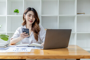 Beautiful Asian woman holding a smartphone and using a laptop, businesswoman texting chatting with partner on laptop scheduling a meeting together. Concept of using technology to communicate.