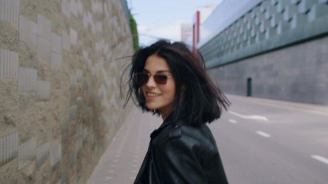 Back view following beautiful seductive happy brunette woman wearing black leather jacket and sunglasses walking along city street turns to camera flirts and smiling on sunny day. Urban style fashion