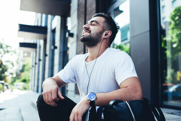 Carefree hipster guy in electronic headphones resting at street urbanity and dreaming during daytime leisure, happy Caucaisan tourist 20s in digital earphones listening audio playlist songs