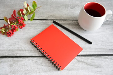 red notebook with black pen and white cup of coffee and branch of barberry with red berries and  autumn leaves on gray wooden background