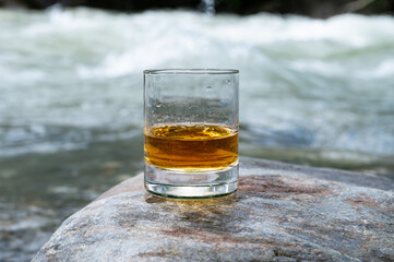 Glass of strong scotch single malt whisky with fast flowing mountain river on background