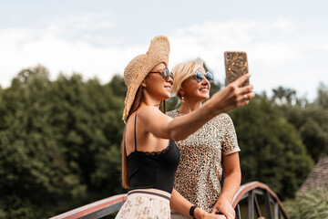 beautiful young woman with a smile and an elderly happy mom in sunglasses in fashionable summer clothes do a joint selfie photo and relax on sunny weekend