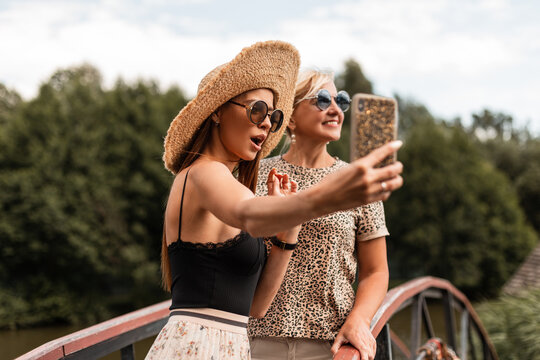 two beautiful funny women happy mom and daughter in fashionable sunglasses with a hat and stylish summer outfit make a joint selfie photo on the smartphone on vacation