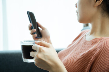 Young Asian woman texting and exploring social media on a smartphone while drinking coffee in morning.