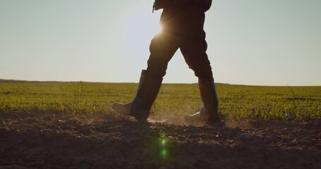 Farmer walks in rubber boots  down a farmer field  dust rising from shoes. Low angle. One part is...