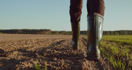 Low angle: man walking in rubber boots in a farmer's field, the blue sky above the horizon. Man walking through an agricultural field. Farmer walks through a plowed field in early spring.