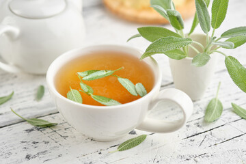 Fresh green sage bunch closeup. Healthy sage herbal tea cup, green leaf of salvia officinalis and tea kettle on white wooden table.