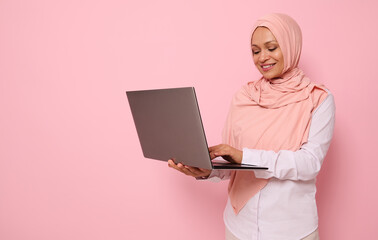 Portrait of a beautiful Muslim Arab woman in pink hijab working on laptop, isolated on colored background with copy space. Successful programmer, IT female worker, start-up, business lady